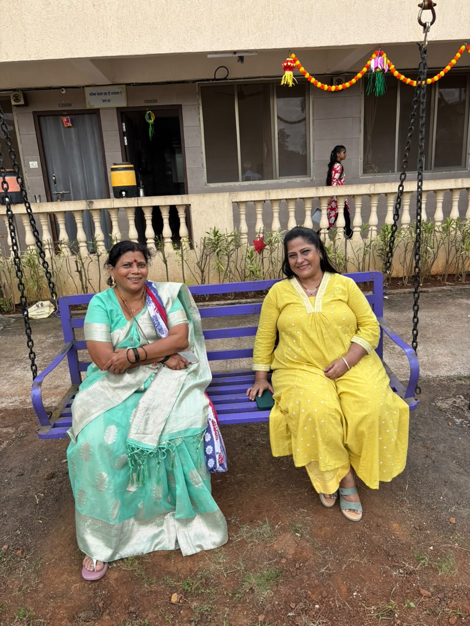 Two women seated together on a garden swing on the campus, with the building behind them